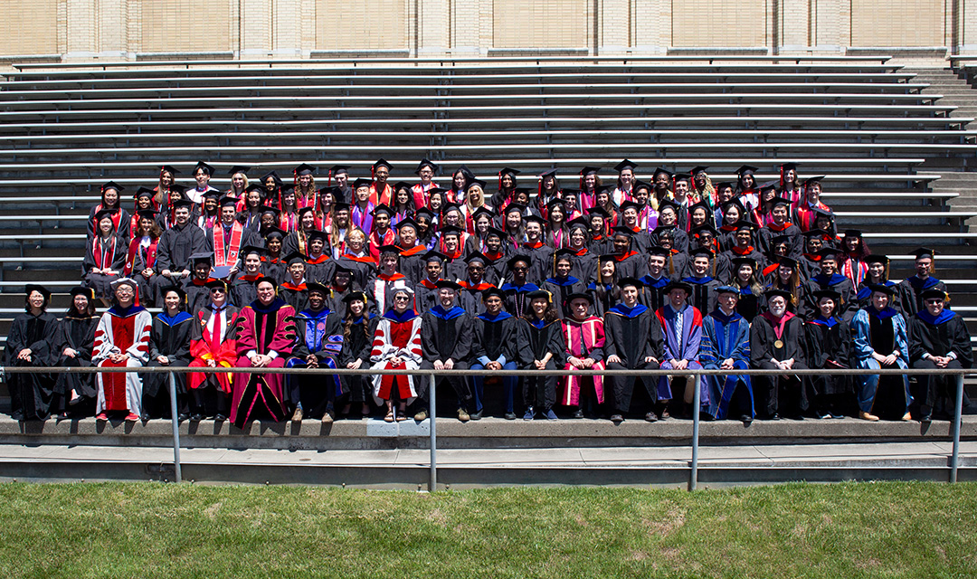 Graduates and faculty wearing regalia sitting outside on bleachers
