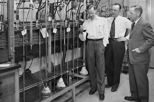 John Speier, Earl Warrick, and Rob McGregor stand in a lab examining a boiling flask containing dark liquid.