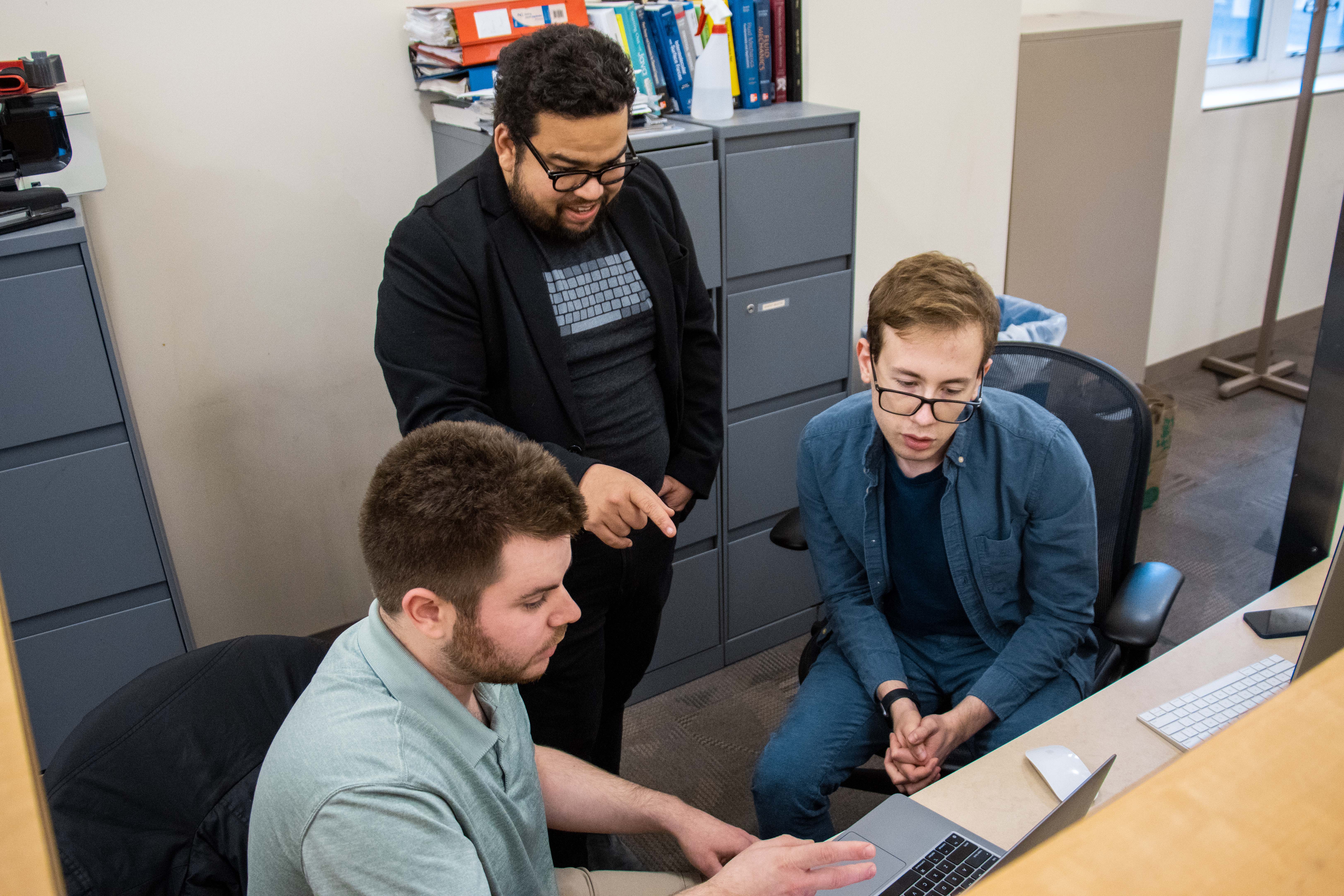 Three men look at one laptop in their research office