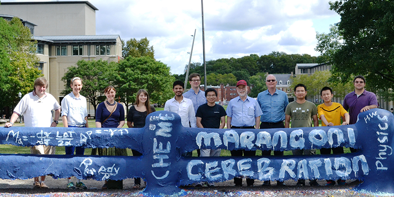  A group of adults pose for a photo behind Carnegie Mellon University’s painted fence, which has the words “ChemE, Physics, Marangoni Cerebration”