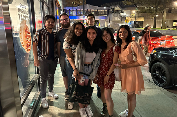 A group of 7 young adults pose for a photo outside a city bar at night.