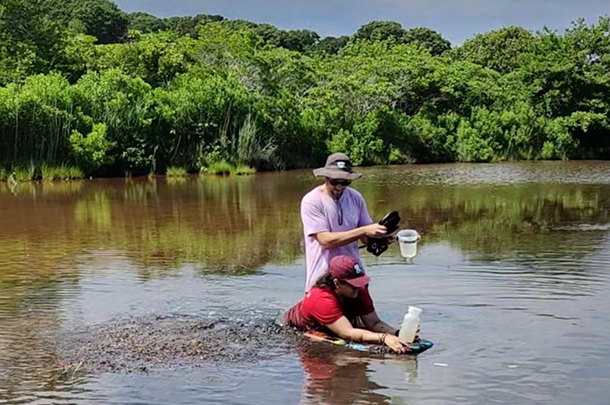 Two adults stand in a shallow river collecting water samples in plastic bottles.