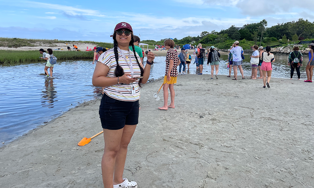 Huda Usman stands on the beach holding a soil sample, with her classmates in the background collecting samples from the salt marsh.