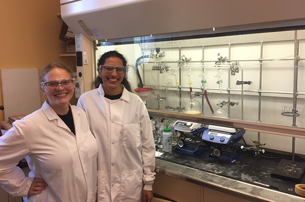 Two women in lab coats and safety glasses standing in front of laboratory equipment