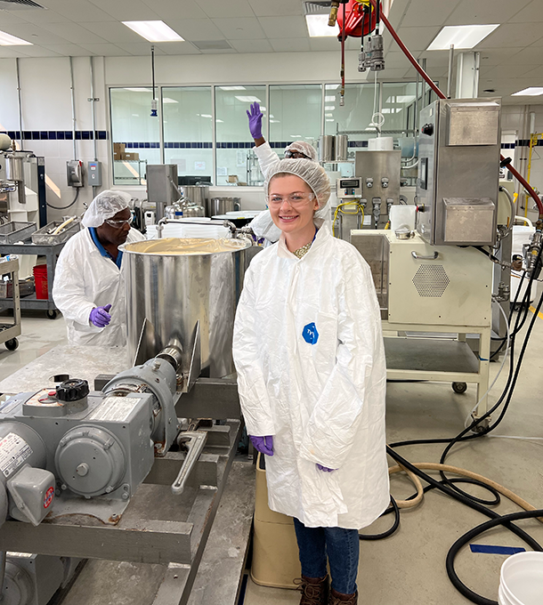 A young woman wearing a white lab coat and hair net stands in an industrial kitchen.