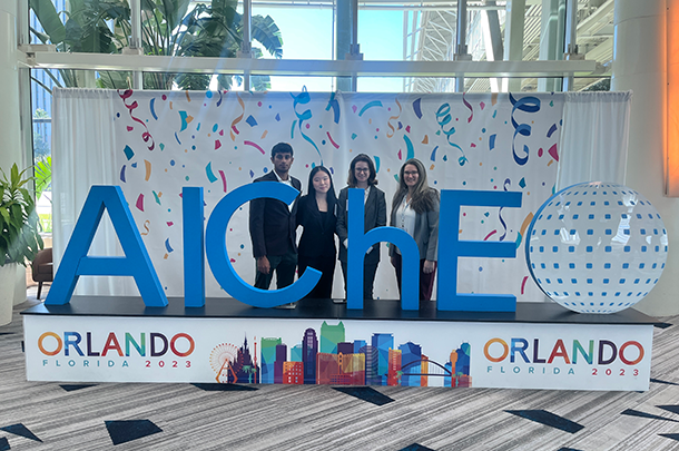 Four adults stand and smile behind a large sign with the AIChE logo.