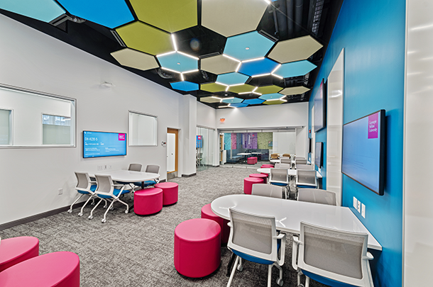 An open room with tables, chairs, and pink ottomans. Both side walls have several LCD screens. Blue, green, and beige hexagon tiles decorate the ceiling.