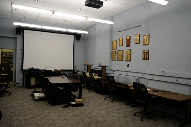 A dim and dull-colored room with tables and chairs and an outdated projector and screen.