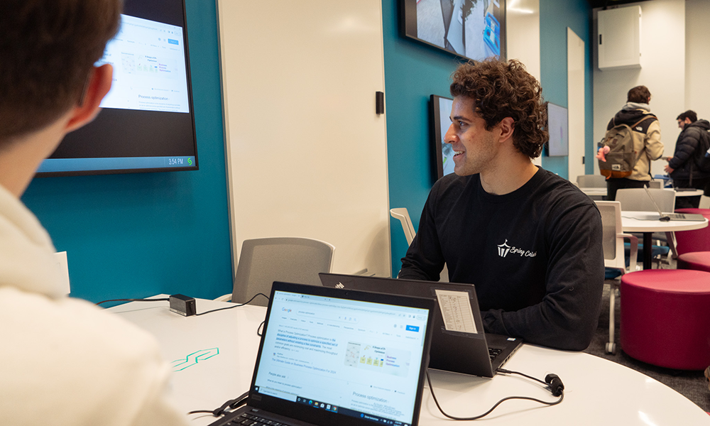 Two students sitting at a table with laptops, looking at display screen on the wall next to them. More tables with students in the background.