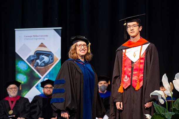 Two adults in regalia stand in front of a row of seated adults wearing regalia