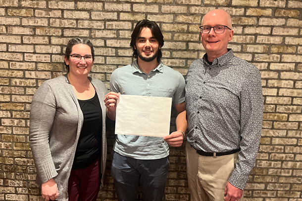 Aleksa Petric stands between two faculty members in front of a brick wall, holding his award certificate