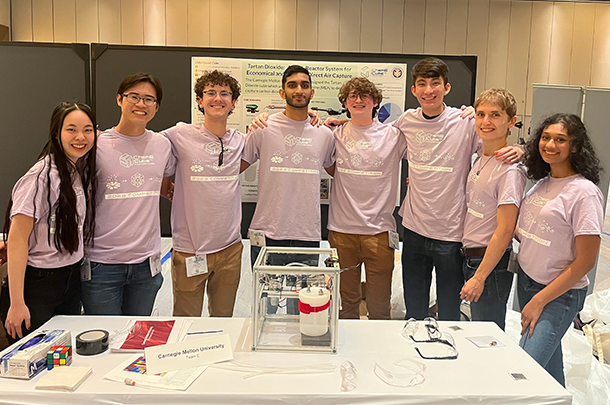 8 students wearing matching ChemE Cube t-shirts stand behind a table on which their cube system is displayed