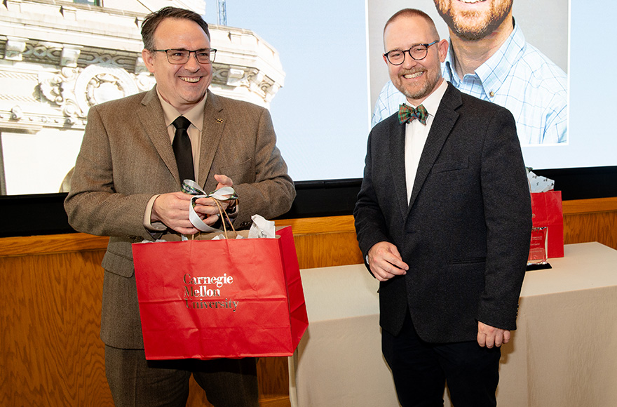 At the awards ceremony, Carl Laird and John Kitchin smile and laugh. Carl is holding a large red gift bag.