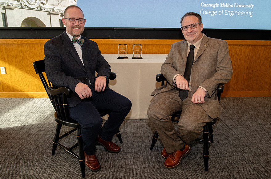 John Kitchin and Carl Laird sit smiling in the captain’s chairs presented during the professorship ceremony. On a table behind them are two rectangular crystal awards.