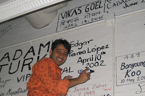 Vikas Goel smiles for the camera while holding a black marker. He is leaning against a white brick wall covered with signatures and dates written in black in different handwriting. His name and the date April 18, 2005 can be seen at the top of the wall.