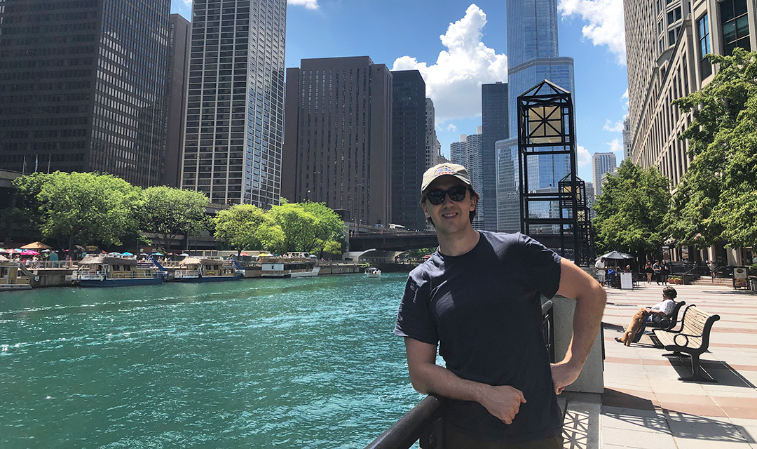 A man wearing sunglasses and a baseball cap smiles while standing next to a river on a sunny day, with a city skyline and bright blue sky in the background.