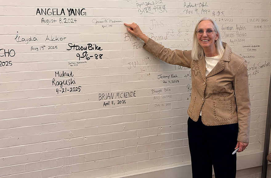 Joanna Livengood stands pointing at a white wall with names and dates written in black marker.