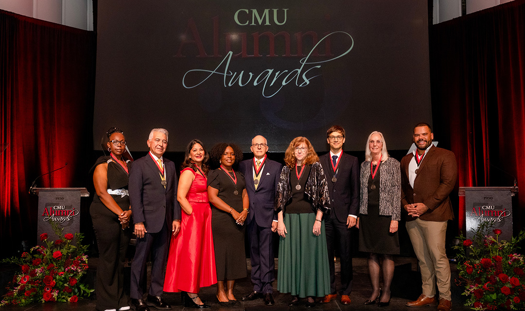 Nine adults wearing suits, dresses, and award medals stand in a line on a stage. The backdrop says C.M.U. Alumni Awards.