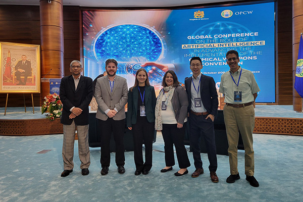 Four adults dressed in business casual attire and wearing nametags stand in a line in front of a large presentation screen.